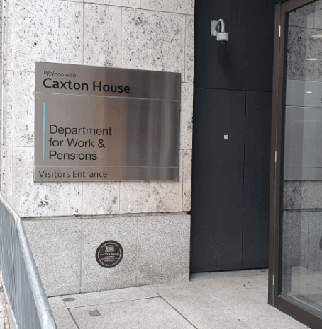 A silver sign on a wall says Welcome to Caxton House, Department for Work and Pensions, Visitors Entrance, with people walking away from the camera on the pavement to the left.