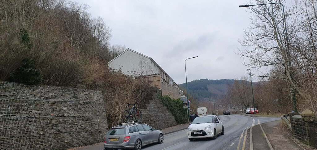 Cars driver along a road with a the tree-covered hillside on the left and a fir-covered hillside in the distance