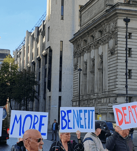 Protesters blocking a street, with the DWP's offices in the background, hold up placards saying 'no more benefit deaths'