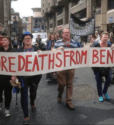 Six people, some of them disabled, carry a banner saying 'No More Deaths from Benefit Cuts' with other disabled people walking behind them along the middle of a street.